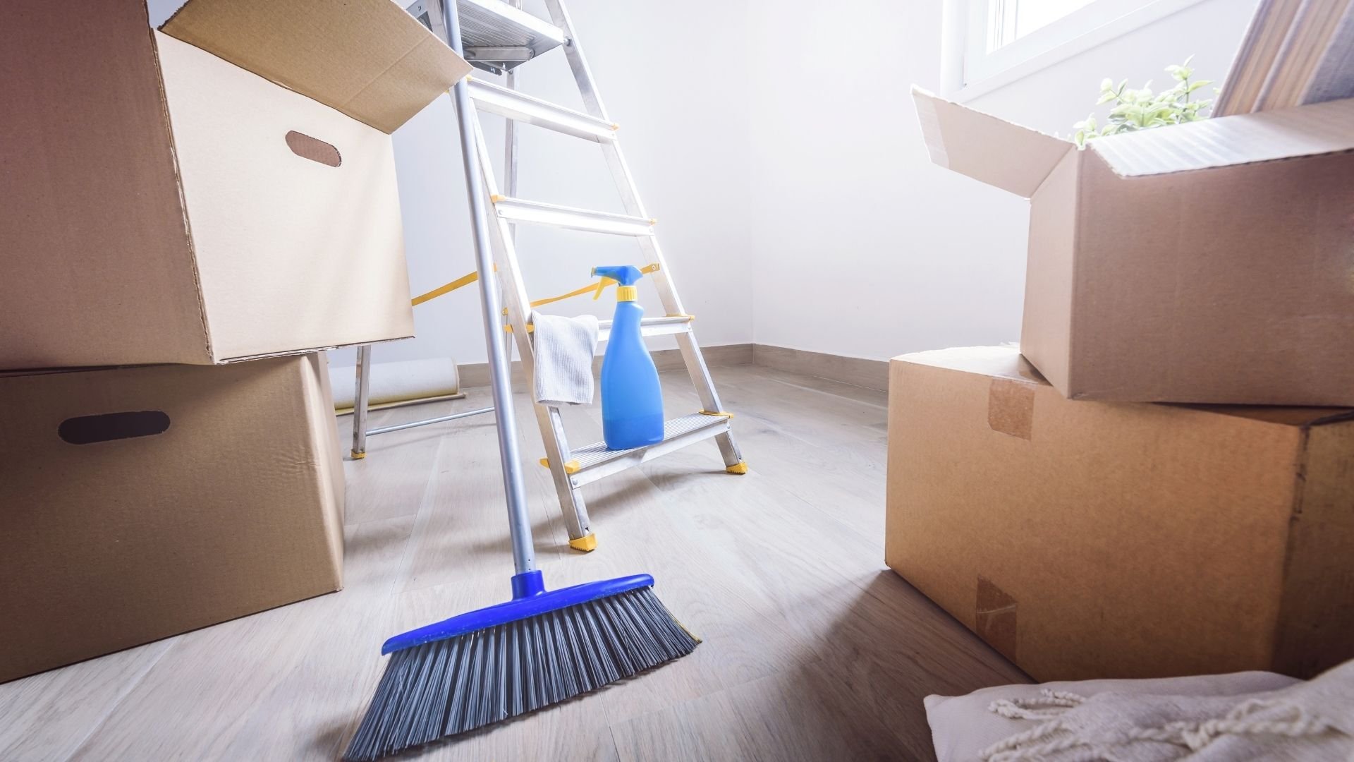 Moving room with cardboard boxes, ladder, broom, and spray bottle for cleaning