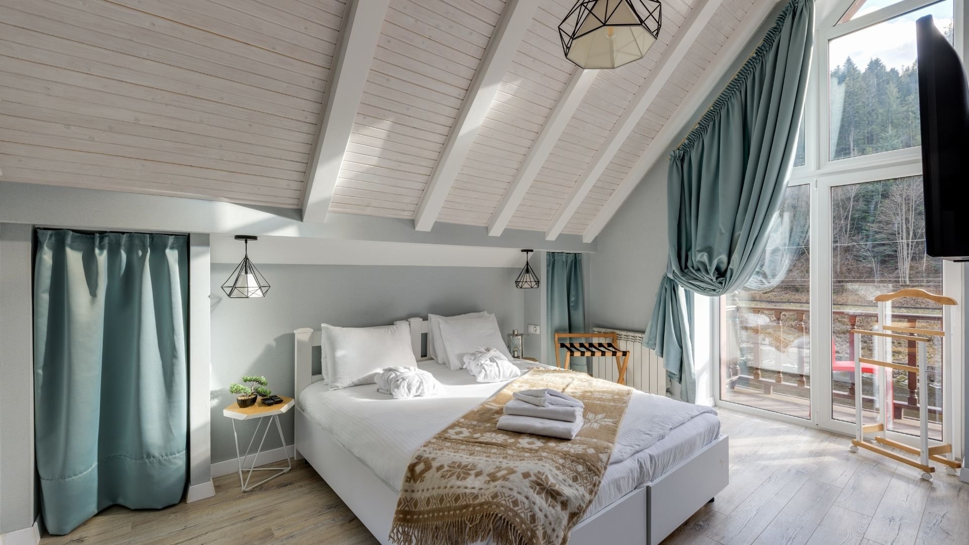 Modern bedroom with vaulted ceiling, queen bed, teal curtains, and large forest views