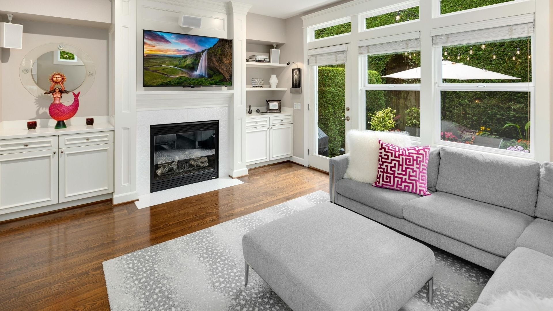 Modern living room with fireplace, gray sofa, large windows overlooking garden, and TV.
