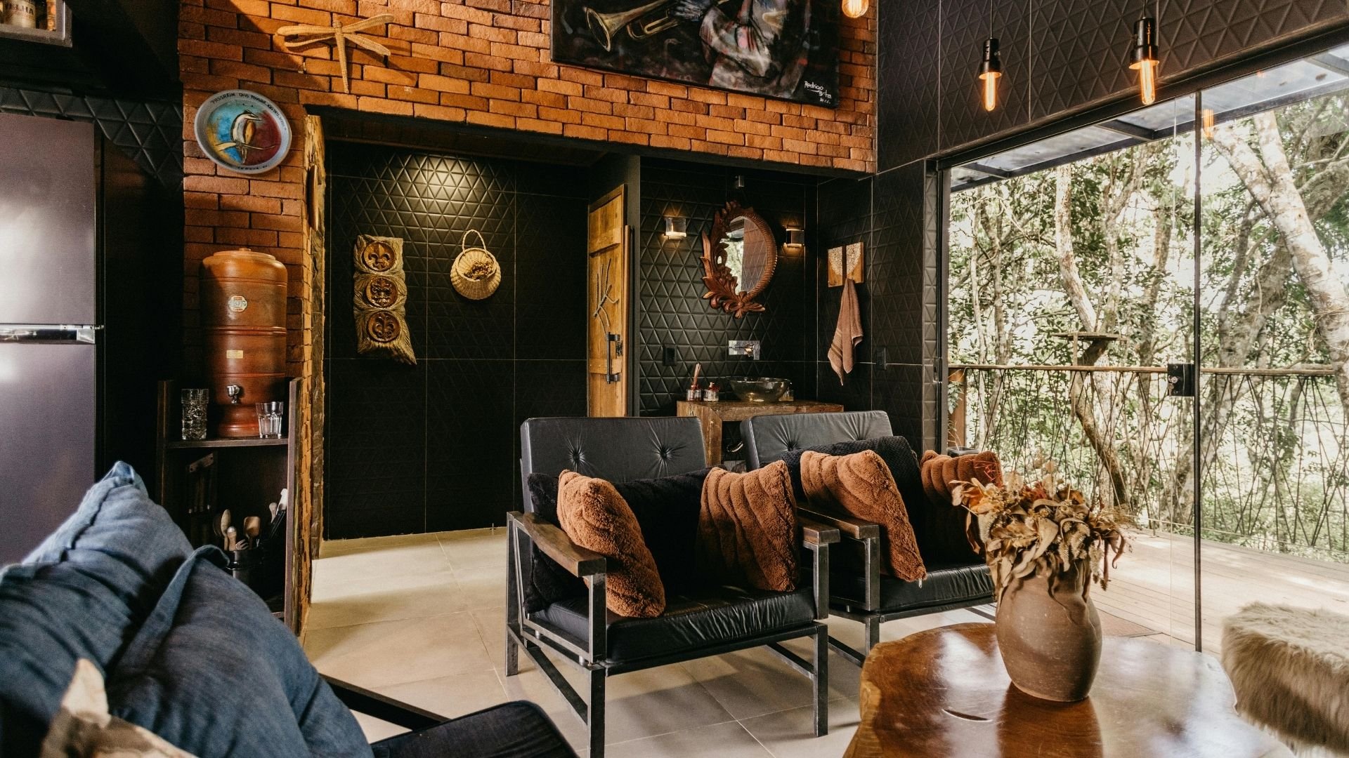Modern living room with black leather seating, brick walls, wooden accents, and glass doors opening to nature.