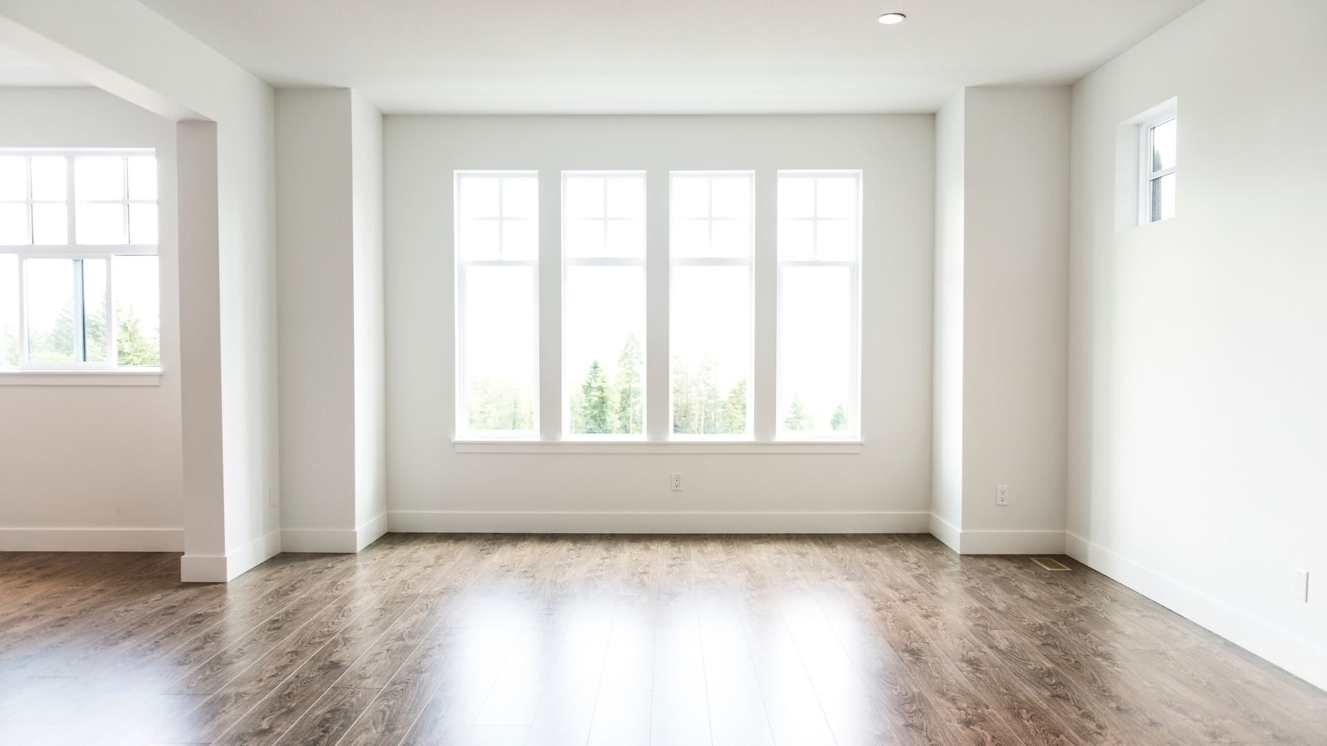 Bright, empty room with wood flooring and four large windows overlooking trees