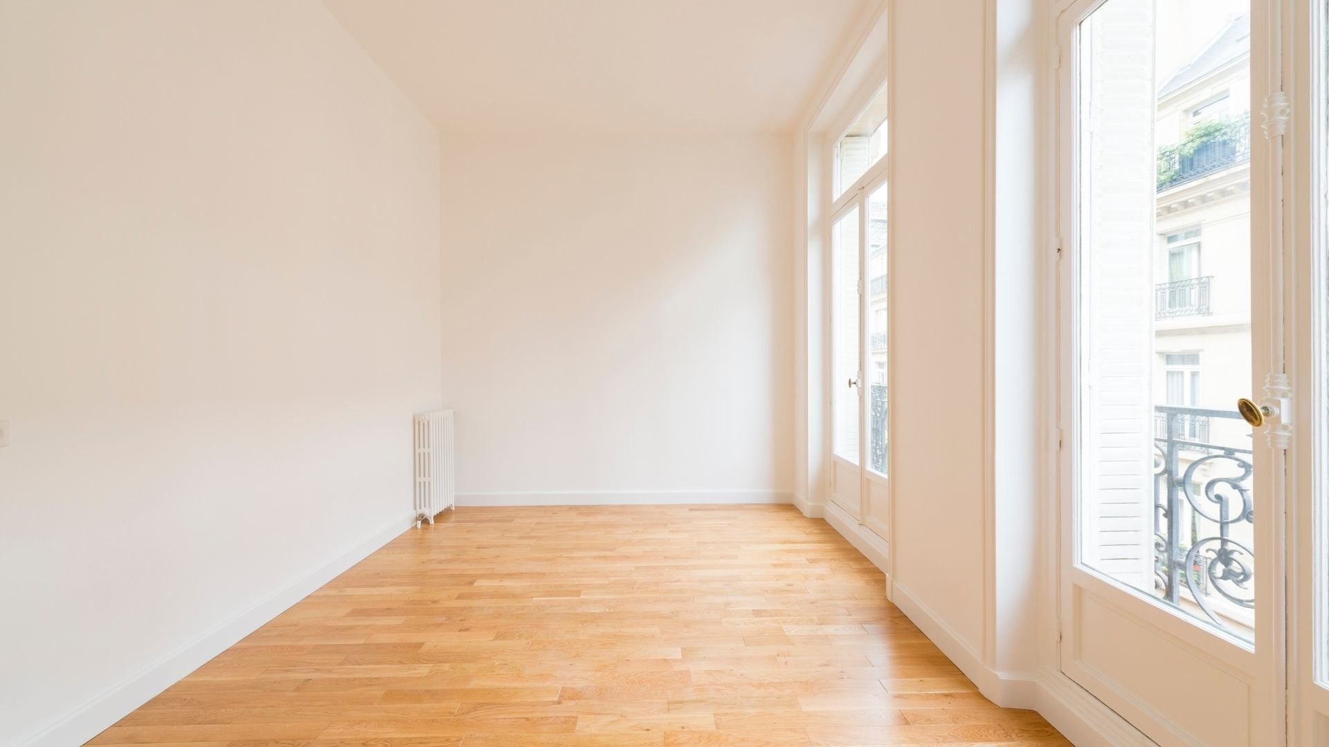 Bright empty room with wooden floor, white walls, and French doors opening to balcony.